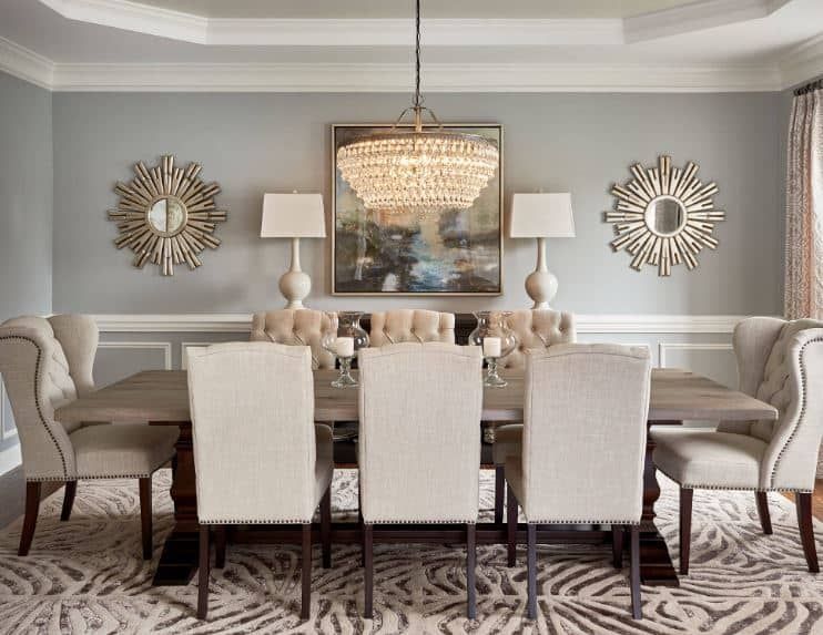 The light gray walls of this formal dining room is adorned with a colorful painting that is flanked by table lamps and a couple of sun-shaped mirrors that pair well…